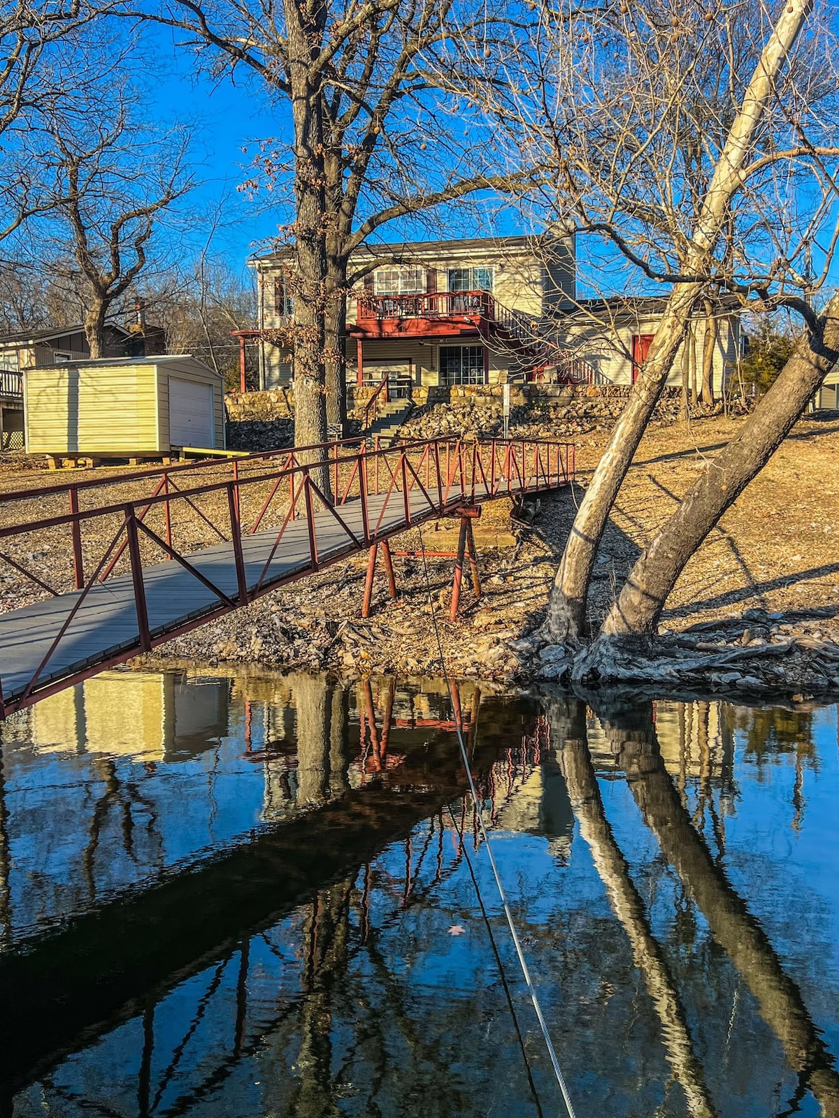 A tranquil lakeside view features a bridge leading to a two-story cottage, reflecting in the calm waters below. Surrounding trees create a natural frame, enhancing the serene atmosphere of the property. The clear blue sky adds to the peaceful setting.