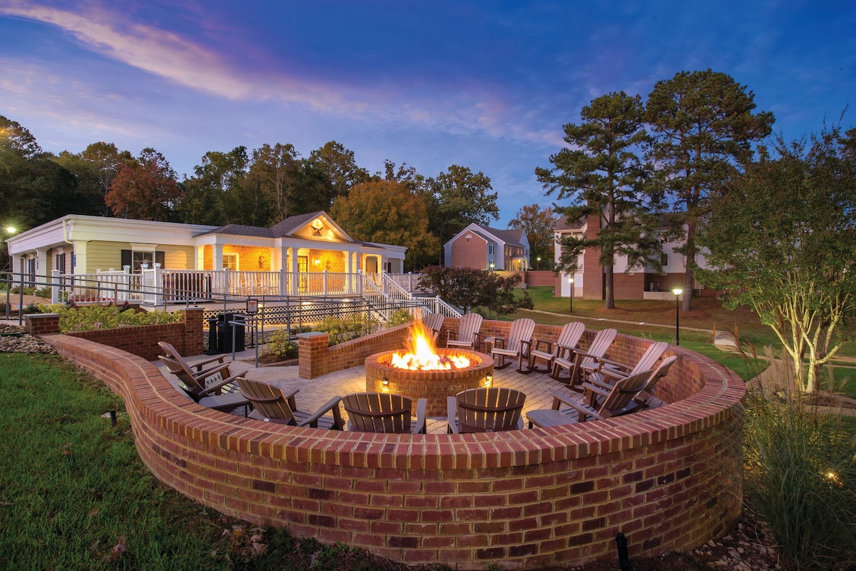 An outdoor fire pit area is surrounded by a circular brick wall, featuring multiple wooden chairs arranged for seating. The building behind is well-lit, showcasing a welcoming entrance. Surrounding trees and grassy areas create a tranquil atmosphere during twilight.