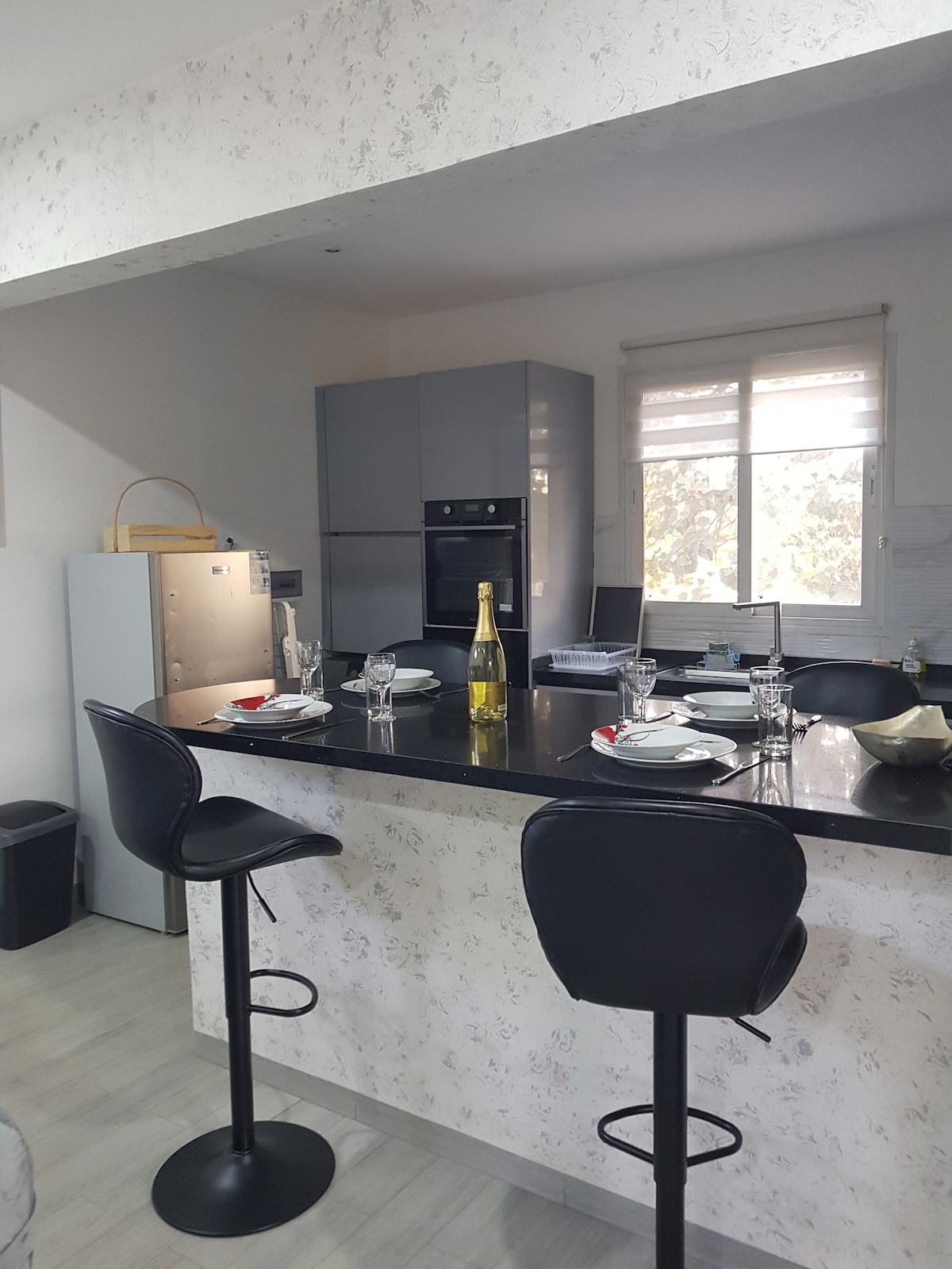 A modern kitchen features sleek gray cabinetry and a black countertop. Two high-backed bar stools are positioned at the counter, with a table set for four. Large windows allow natural light to brighten the space, complemented by a stainless steel refrigerator and built-in oven.