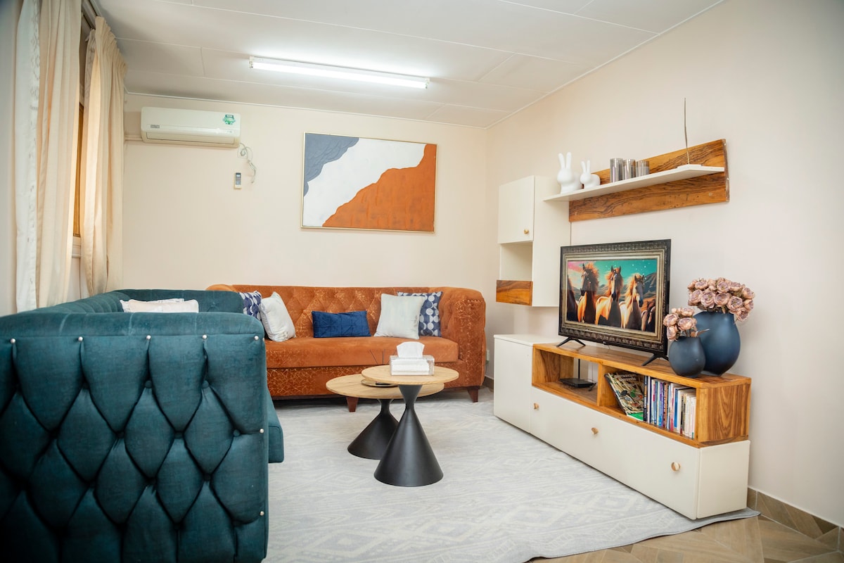 A spacious living area is furnished with a blue tufted couch and a brown leather sofa. A circular coffee table is centered on a light rug, while a TV stand displays a television and decorative items. Natural light enters through sheer curtains.