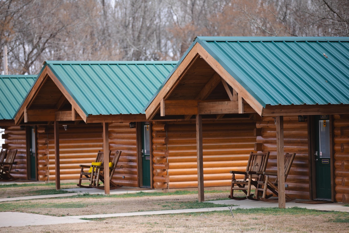 A row of luxury log cabins is shown with distinctive green metal roofs. Rocking chairs are placed on the porches, inviting relaxation. The well-maintained ground features a pathway leading to each cabin's entrance, framed by natural greenery in the background.