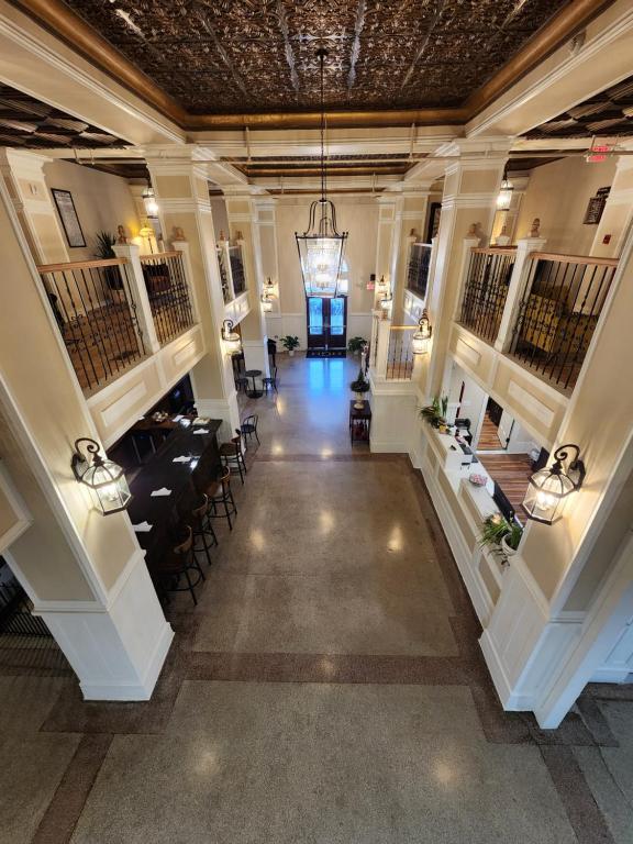 A grand lobby showcases an elegant design with high ceilings and ornate lighting fixtures. A long bar runs along one side, complemented by comfortable seating. Indoor plants are positioned throughout, adding a touch of greenery. The mezzanine level provides additional seating, enhancing the space's inviting atmosphere.