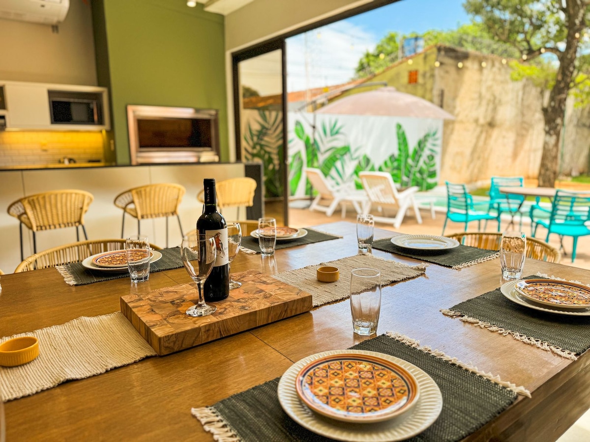 A dining area featuring a wooden table set with plates and glasses, accompanied by a bottle of wine in the center. Large glass doors open to a patio with a private pool and lounge chairs, surrounded by greenery, enhancing the inviting atmosphere of the space.