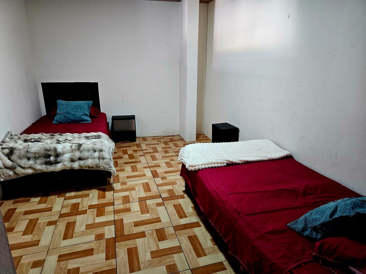 A spacious room features two beds covered with red bedspreads and accented by soft throws. Nightstands are positioned beside each bed. The flooring consists of a patterned wood design, contributing to a cozy ambiance in the bright, uncluttered space.