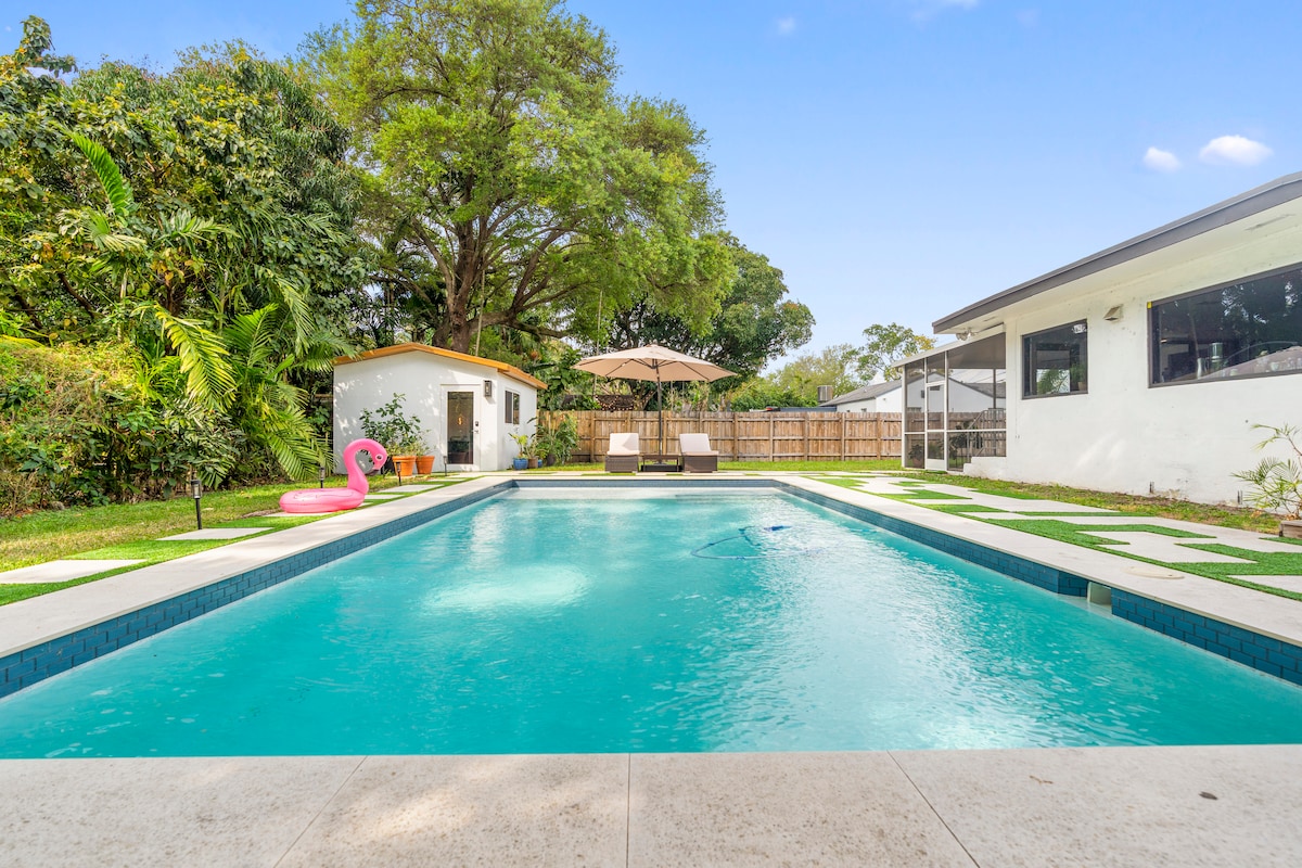 A tranquil pool area is featured, showcasing a sparkling blue pool surrounded by lush greenery. Two lounge chairs with a large umbrella provide shaded seating. A small white cottage and a pink inflatable float add a playful element to the serene outdoor space.