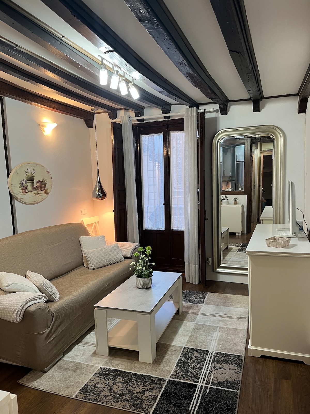 A cozy living area is presented with a light-colored sofa and a simple coffee table atop a patterned rug. Natural light enters through large windows, and a full-length mirror reflects the space. Wooden beams and warm lighting create an inviting ambiance.