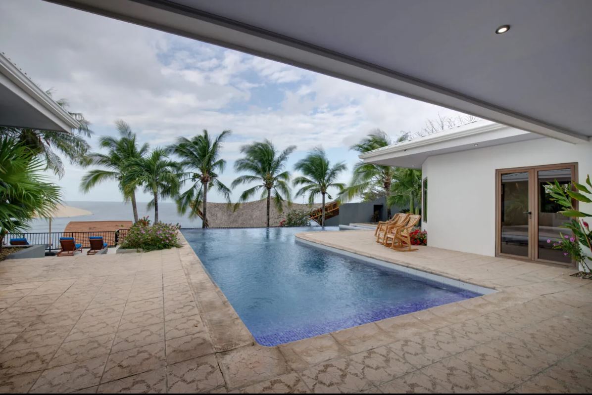 An expansive oceanfront infinity pool is depicted, surrounded by swaying coconut palms. A terrace features several loungers, with a view overlooking the water. The villa's modern architecture is visible, complementing the scenic outdoor space.