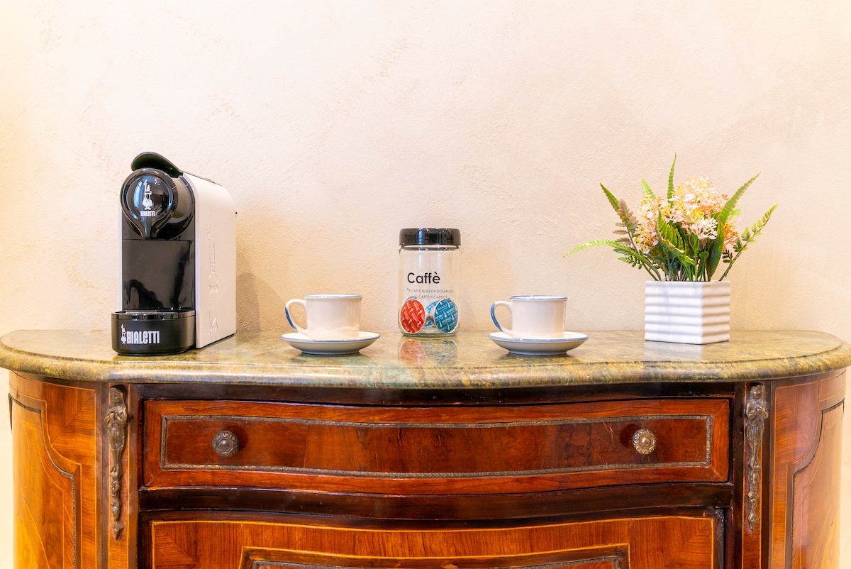 A sleek coffee machine is positioned on a polished countertop beside two white cups. A jar labeled 'Caffè' sits centrally, complemented by a small potted plant in a white container. The wooden console adds a classic touch to the arrangement.