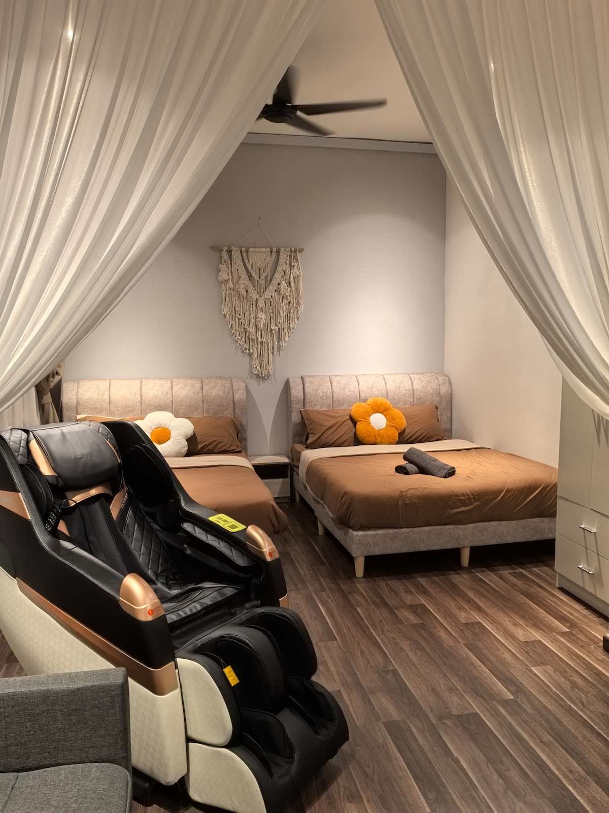 A sleeping area features two spacious beds dressed in neutral-toned linens. A massaging chair is positioned nearby. Soft curtains frame the space, contributing to a cozy ambiance, while a decorative wall hanging adds an artistic touch.