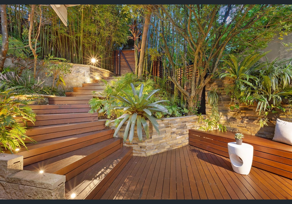 A serene outdoor space features wooden steps leading through lush greenery and thoughtfully arranged plants. Soft lighting enhances the natural beauty, illuminating the pathway and creating a peaceful retreat. Stone walls provide texture, while landscaped areas invite relaxation among the vibrant foliage.