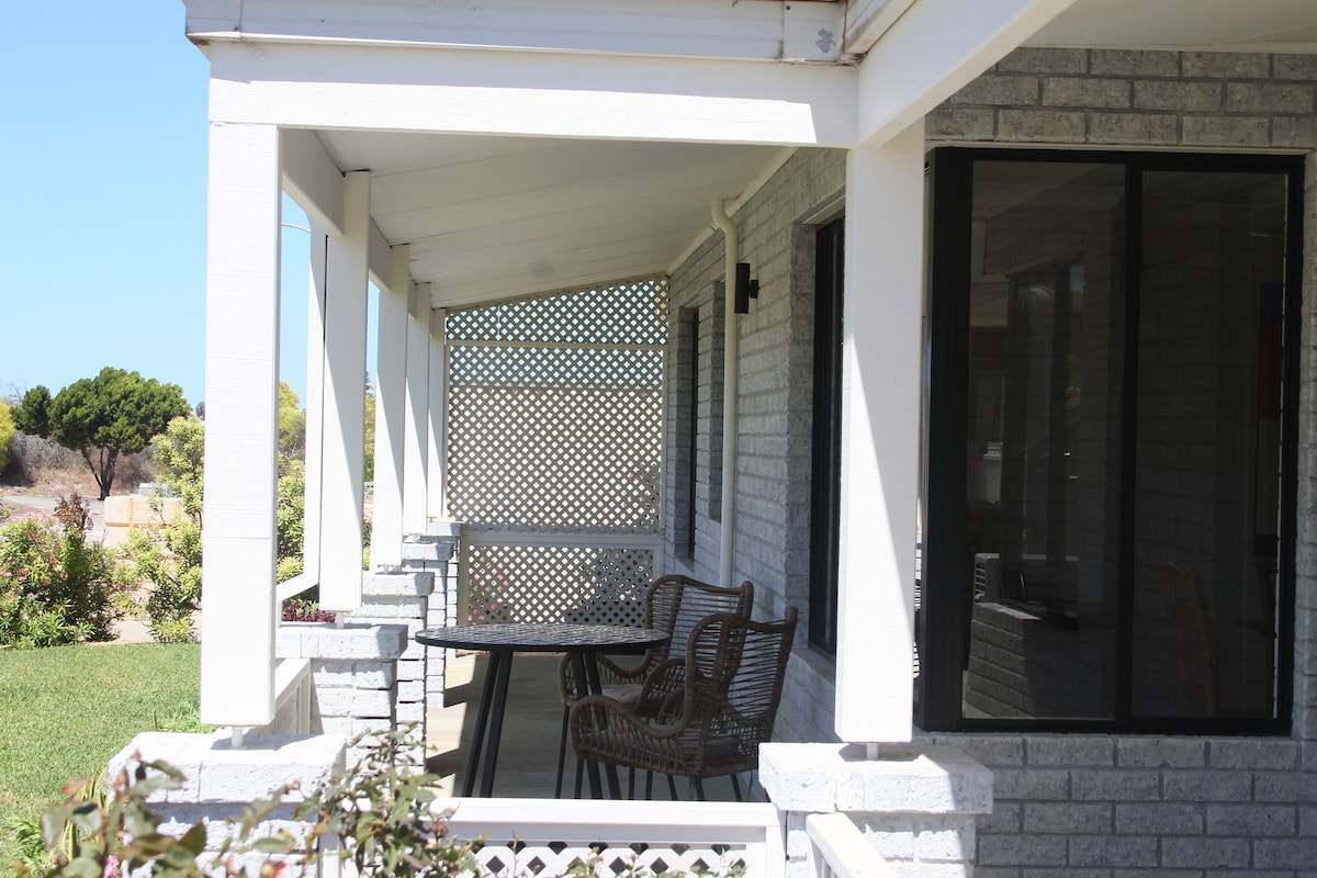 A covered outdoor patio area is visible, featuring a table and multiple wicker chairs. Light filters through the latticework, providing a shaded spot for relaxation. The surrounding grassy area offers a serene view, enhancing the outdoor experience.