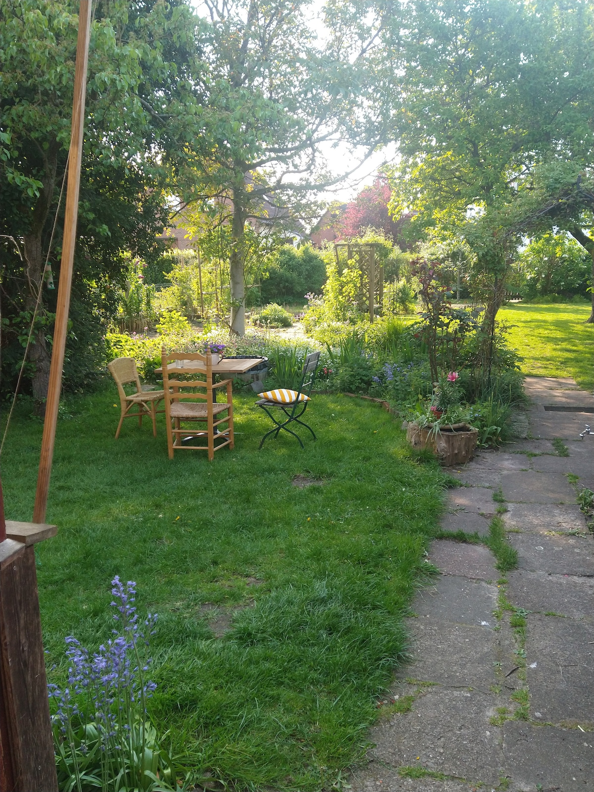 A serene garden area is visible, featuring a grassy lawn with a stone pathway leading to a wooden table and chairs. Various plants, flowers, and trees surround the space, creating a relaxed outdoor setting with dappled sunlight filtering through the foliage.