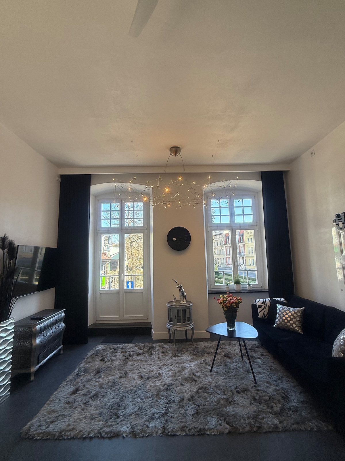 A spacious living area is illuminated by natural light coming through large windows adorned with dark curtains. A comfortable sofa is positioned next to a round coffee table. Decorative accents, including flowers and cushions, create a welcoming ambiance.