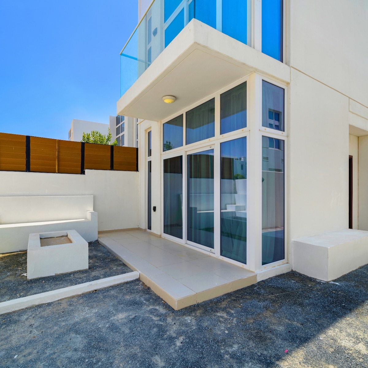 An outdoor terrace features light-colored tiles and a built-in seating area. Large glass doors provide access to the interior. The surrounding area is gravel, and the backdrop contains a combination of wooden fencing and soft greenery, creating a serene outdoor space.