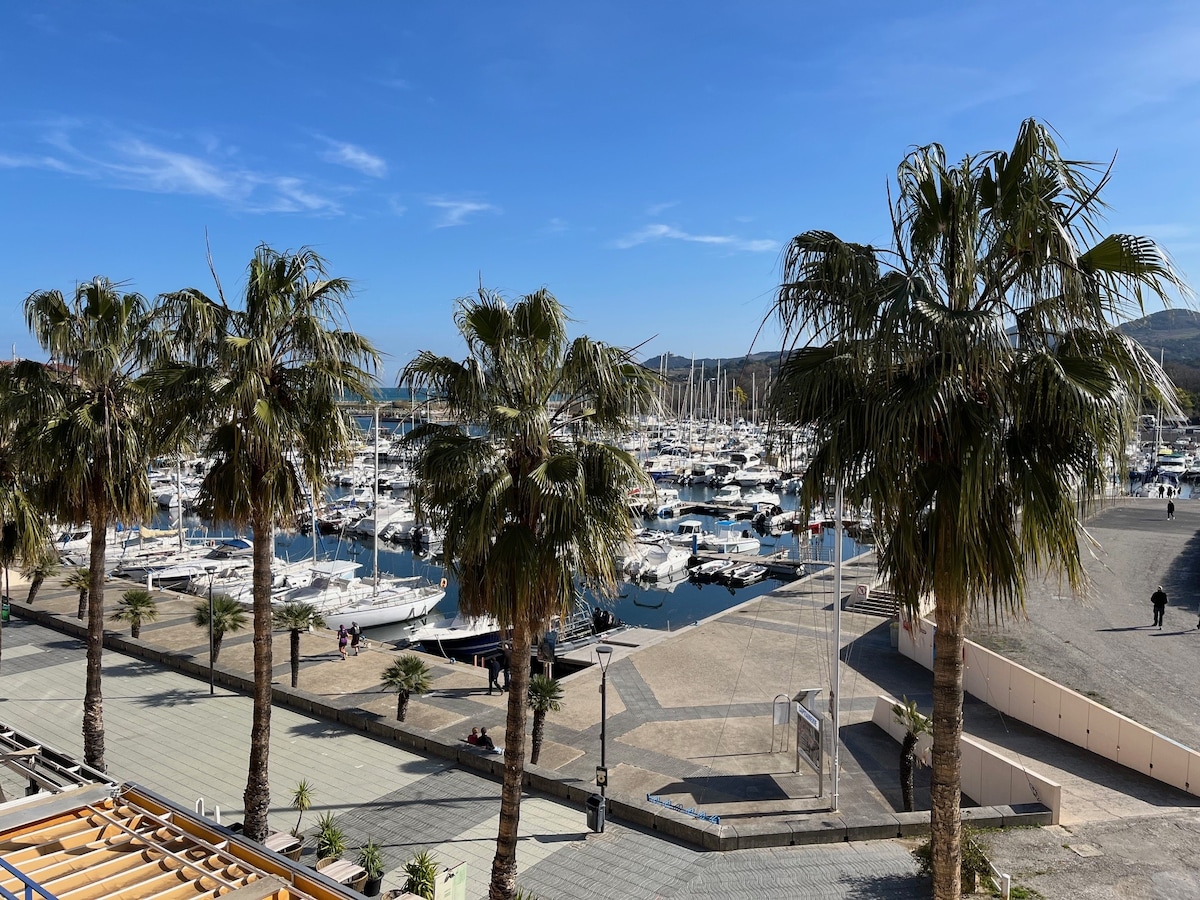 A view of a vibrant marina filled with numerous boats, framed by palm trees. The sunny sky enhances the serene atmosphere, while the waterfront area features a wide walkway and glimpses of nearby buildings. The scene reflects a peaceful day at the port.