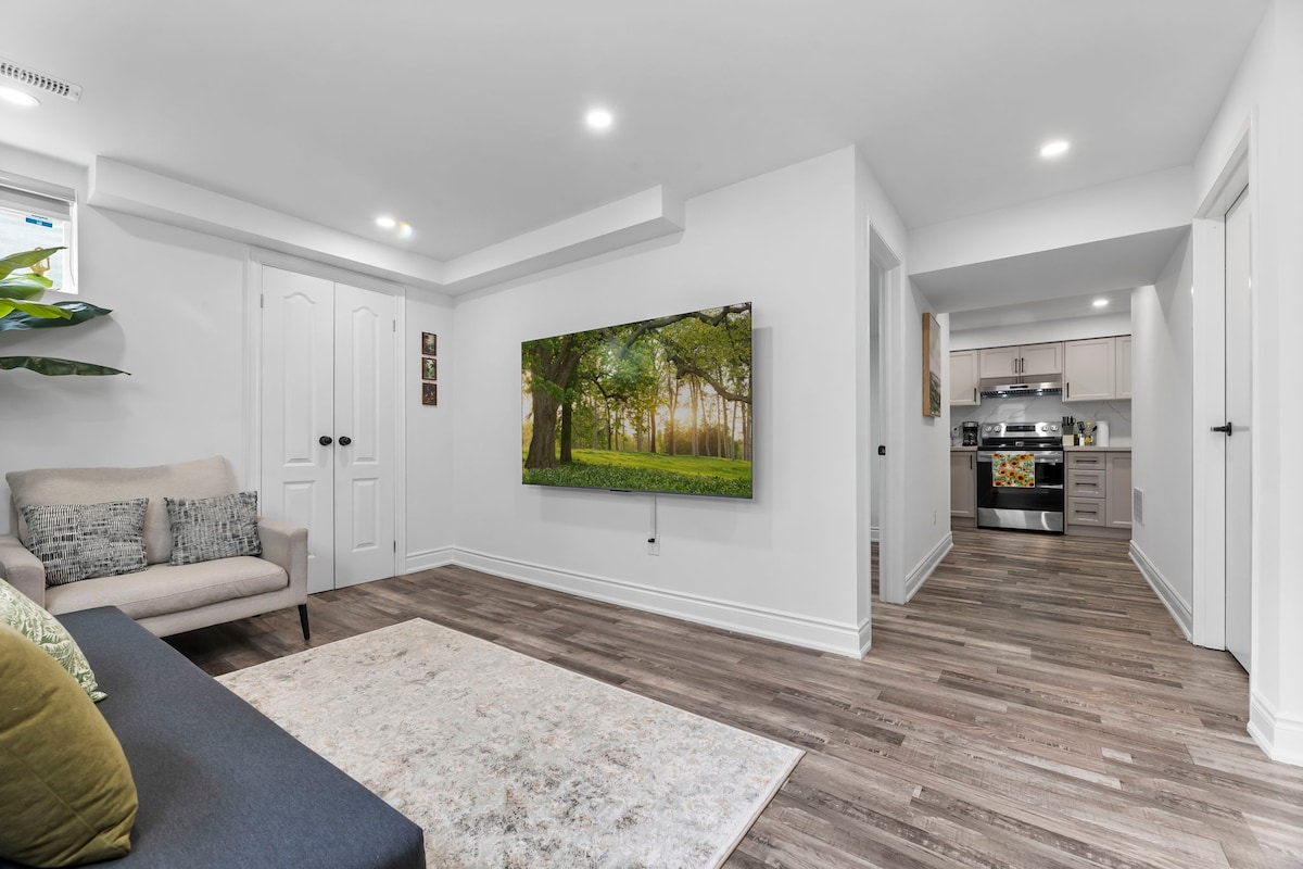 A cozy living area is showcased, featuring a modern sofa and a spacious rug. A large wall-mounted Smart TV displays a serene forest scene, while a doorway leads to a fully equipped kitchen in the background, enhancing the inviting atmosphere.