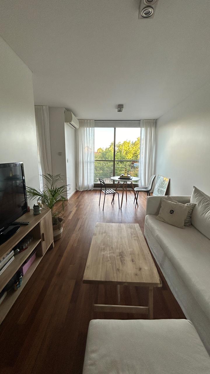 A bright living space features a comfortable sofa and a wooden coffee table. Large windows are draped with sheer curtains, allowing natural light to illuminate the room. Outdoor greenery is visible through the glass, enhancing the tranquil atmosphere.