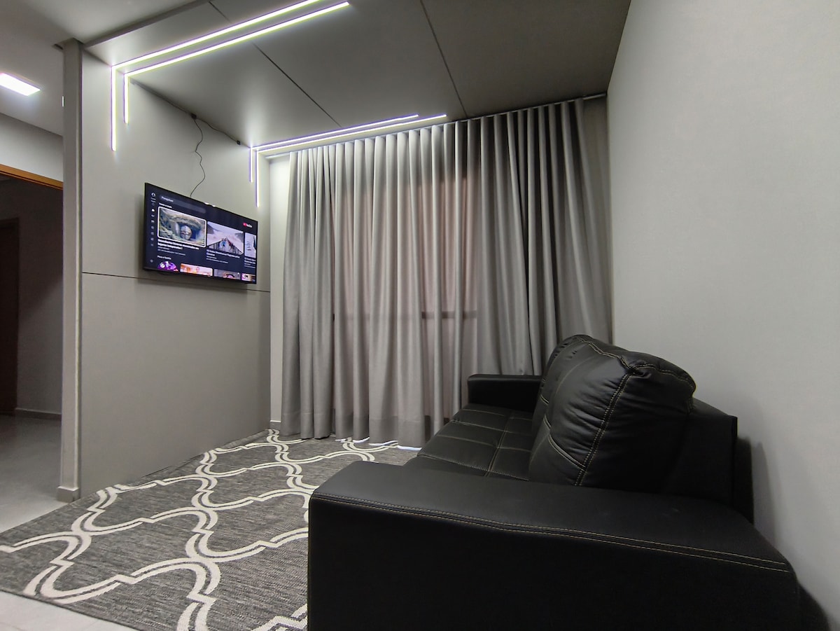 A comfortable seating area features a dark leather sofa positioned against a light gray wall. A television is mounted on the wall opposite a large window, which is dressed with sheer curtains. A patterned area rug adds texture to the space.