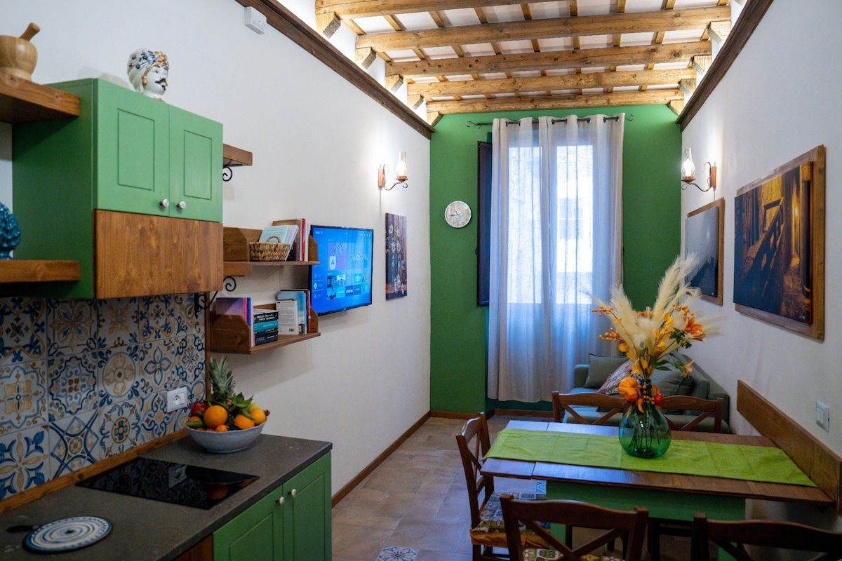 A functional living area features a green kitchen with open shelving and decorative elements. A wooden dining table is set for four, accompanied by a vase of dried flowers. Natural light filters through sheer curtains, illuminating the walls adorned with various artworks.