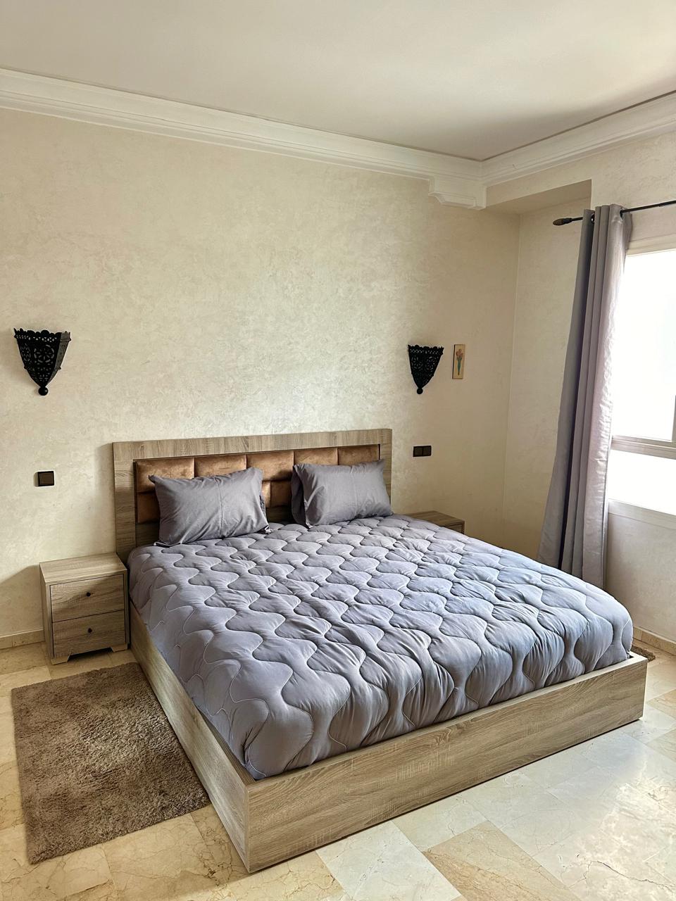 A well-appointed bedroom features a spacious bed adorned with a light gray comforter. Two bedside tables are positioned on either side, while wall sconces add subtle illumination. Natural light filters through a nearby window, with soft gray curtains enhancing the serene atmosphere.