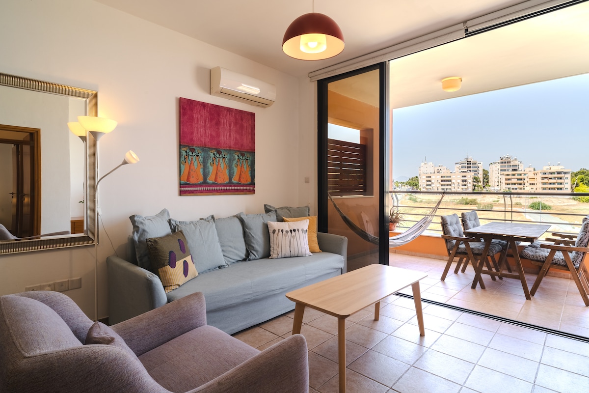 A living area is shown with a gray sofa and an accent chair positioned around a light wood coffee table. A large sliding door opens to a terrace with outdoor seating. A colorful wall art piece and a floor lamp enhance the space, while neutral tiles provide a clean foundation.