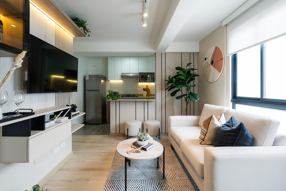 A modern living space is depicted, featuring a light-colored sofa and a round coffee table. A large television is mounted on the wall, complemented by built-in shelving. The kitchen area is visible in the background, showcasing sleek cabinetry and appliances, while greenery adds a touch of nature.