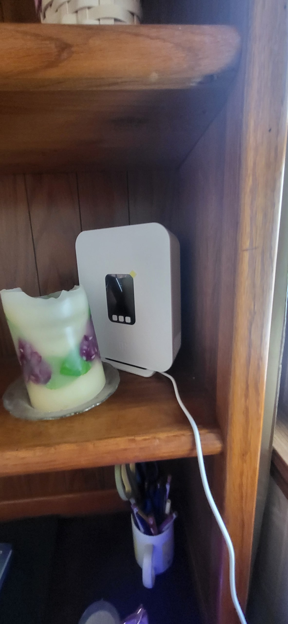 A close-up view of a wooden shelf featuring a partially melted candle with purple accents and a white device plugged into the wall. The background highlights the warm tones of the wood and the soft natural light coming through the window.