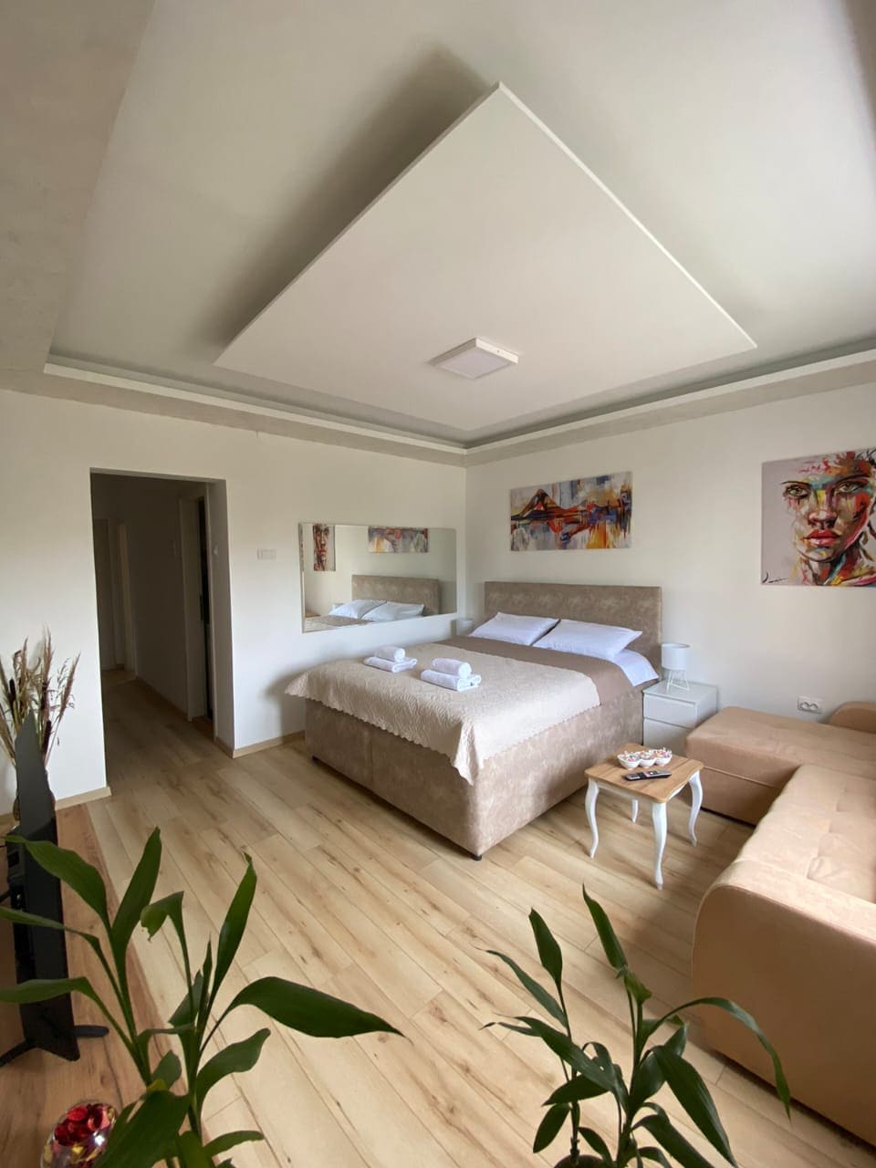 A spacious studio features a large bed with soft linens, positioned against a neutral-toned wall. Natural light filters through the window, illuminating the light-colored wooden floors. A cozy seating area includes a light-colored couch, while decorative elements add a touch of warmth.