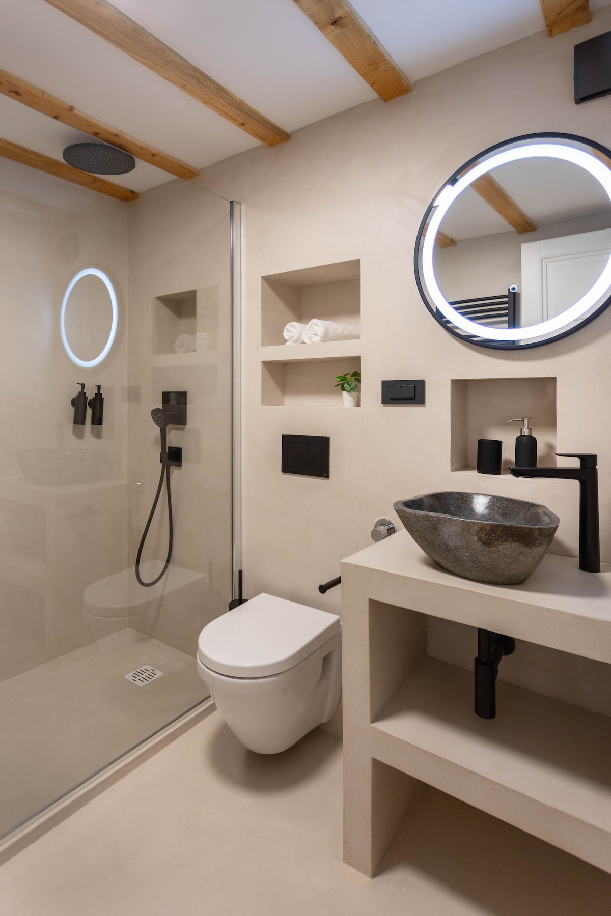 A contemporary bathroom is featured with a walk-in shower enclosed by glass panels. A spacious vanity holds a modern basin and faucet, while shelves display fresh towels and a small plant. A circular mirror with integrated lighting adds a sleek touch.