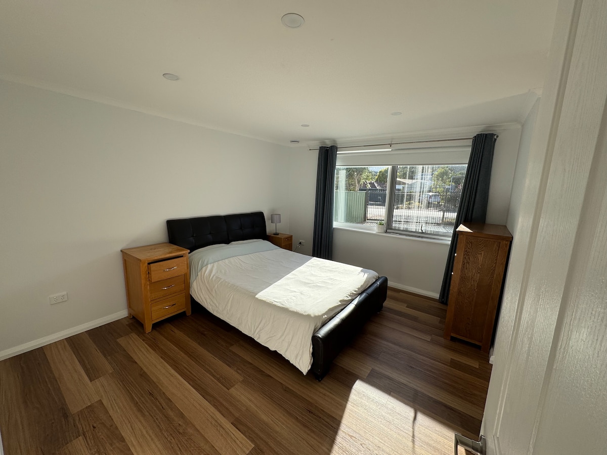 A light-filled bedroom features a spacious bed with a dark leather headboard, accompanied by two wooden nightstands. A large window with curtains allows natural light to brighten the space, while warm wooden flooring adds to the overall ambiance.