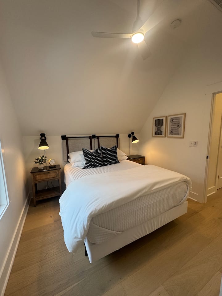 Cama tamaño queen con mesitas de noche, luces de pared y ventilador de techo. Ropa de cama lujosa y persianas celulares opacas.
