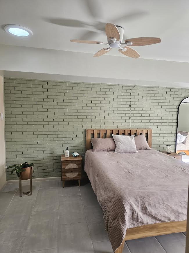 The sleeping area features a spacious bed against a green-brick wall. A nightstand with a decorative plant is positioned beside the bed. A ceiling fan with wooden blades is visible, and a full-length mirror reflects elements of the room.