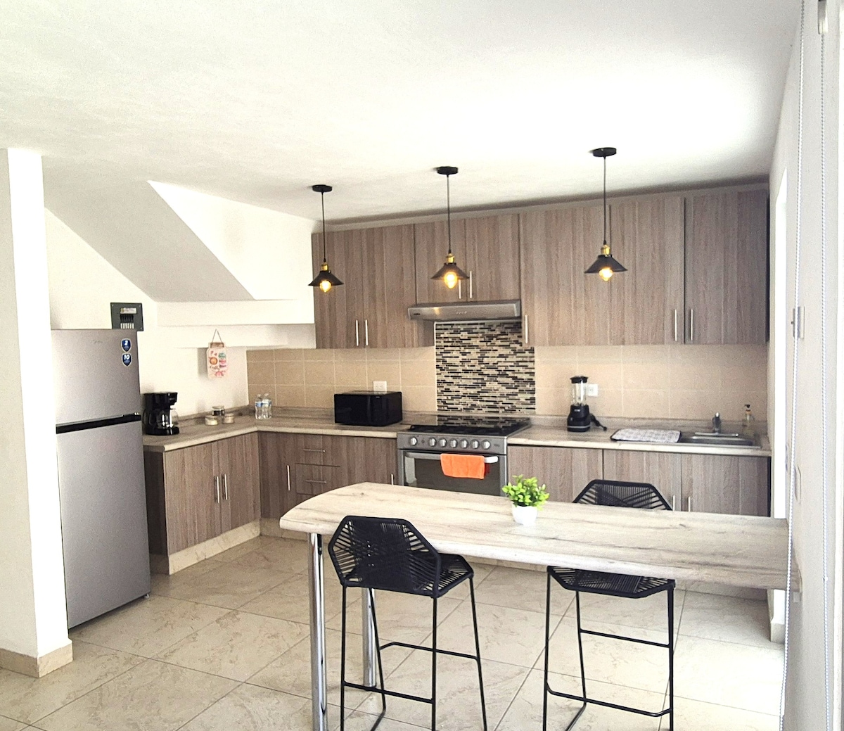 A modern kitchen is equipped with essential appliances, including a refrigerator, oven, and coffee maker. Practical cabinets are complemented by a textured backsplash. A dining table with two bar stools is situated nearby, providing a comfortable space for meals.