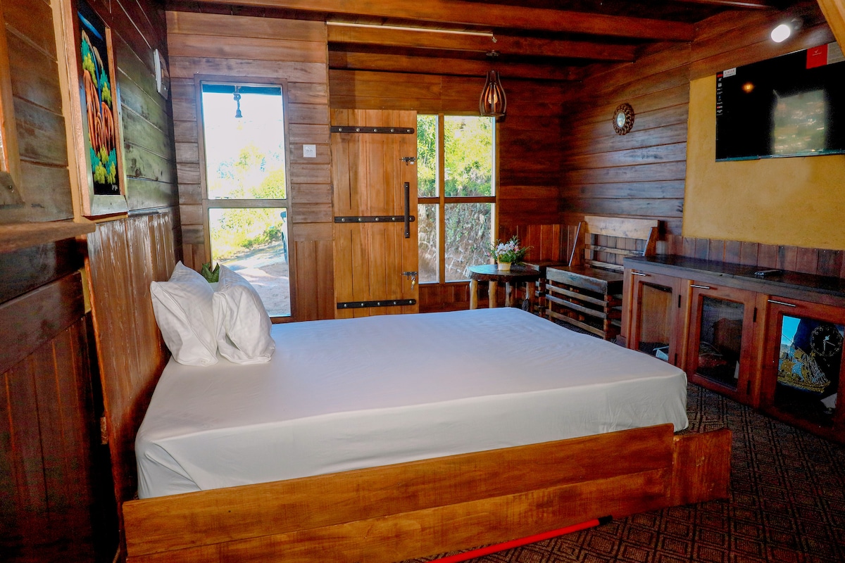 A cozy sleeping area features a spacious bed with crisp white linens, surrounded by warm wooden walls. Natural light filters in through a large window, and a rustic wooden door adds charm. A small seating area includes a table and chairs, enhancing the room's inviting nature.