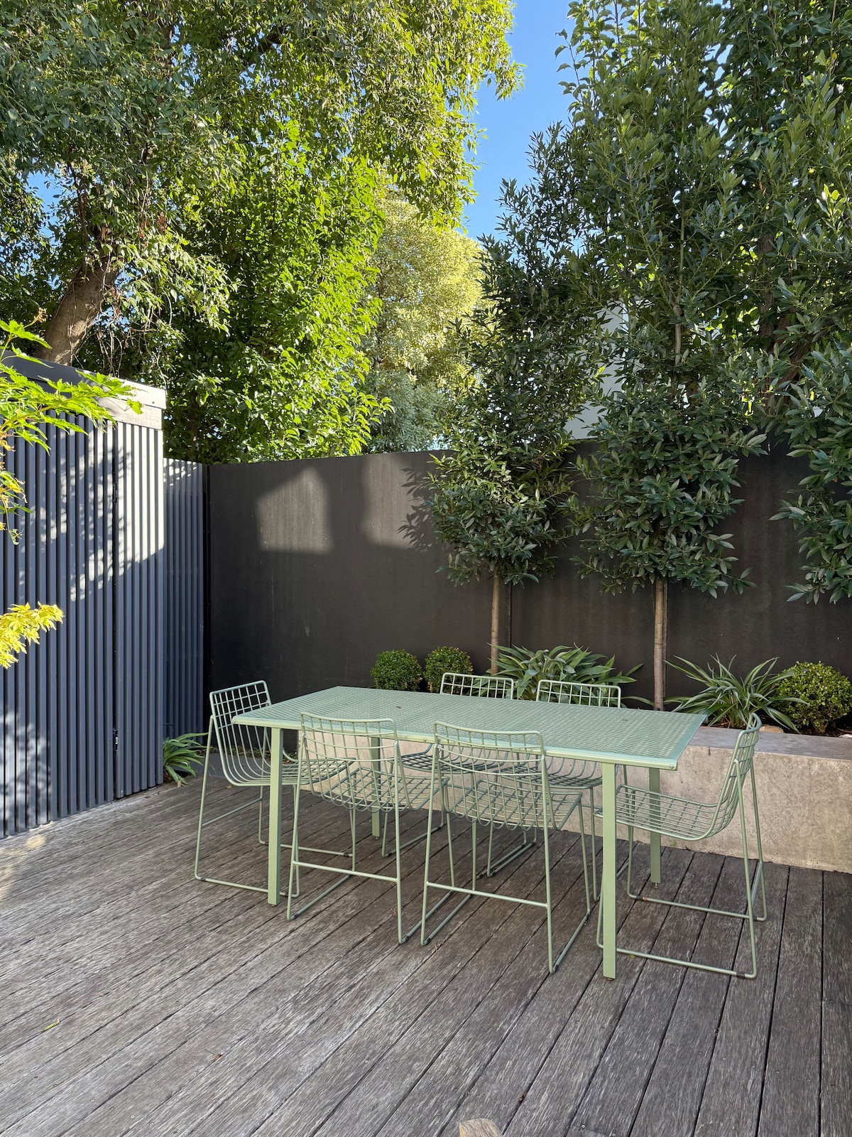 An outdoor dining area features a rectangular table surrounded by six wire chairs. Lush greenery and potted plants create a natural backdrop, while textured wooden decking adds warmth to the space. A tall wall provides privacy, enhancing a peaceful setting.