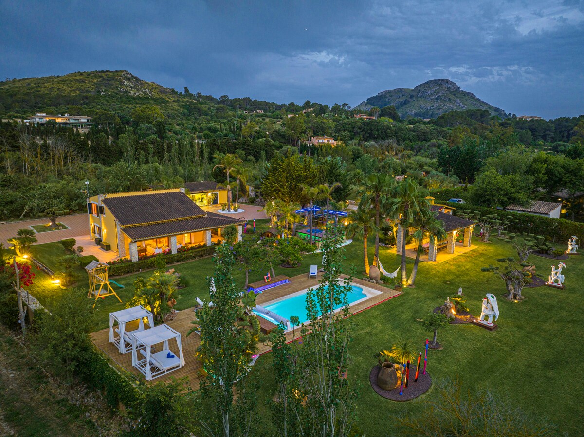 An aerial view captures a spacious property surrounded by greenery, featuring a manicured lawn, illuminated pool, and various sculptures. The villa is complemented by outdoor lounging areas and palm trees, all set against a backdrop of hills under a cloudy sky.