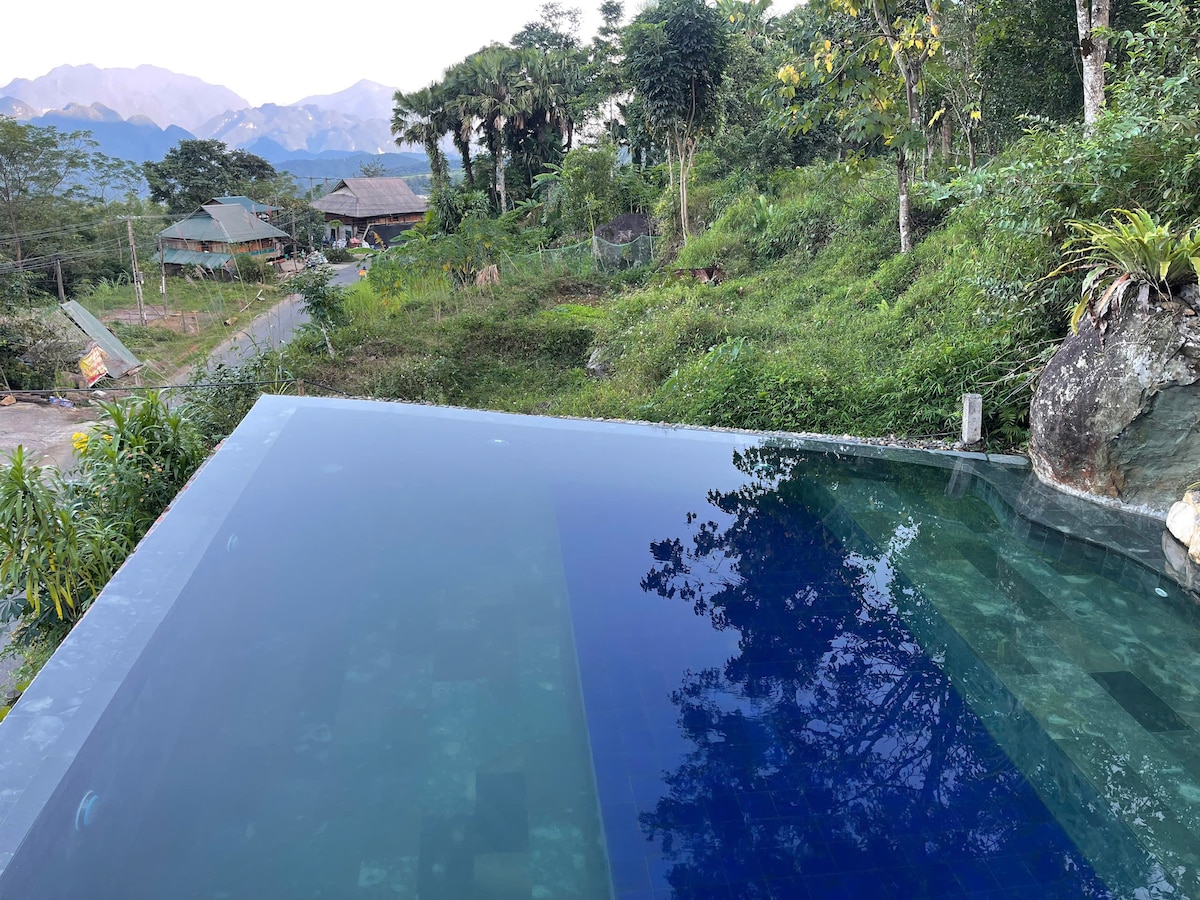 An infinity pool is positioned at the edge of a hillside, reflecting the surrounding greenery and distant mountains. The serene water blends seamlessly with the horizon, creating a tranquil ambiance amidst nature.