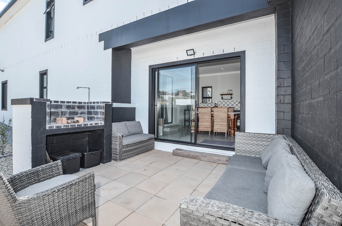 An outdoor seating area features a spacious patio with comfortable grey wicker sofas positioned around a stone surface. A built-in braai area complements the setting, and large sliding glass doors provide access to the interior dining space.