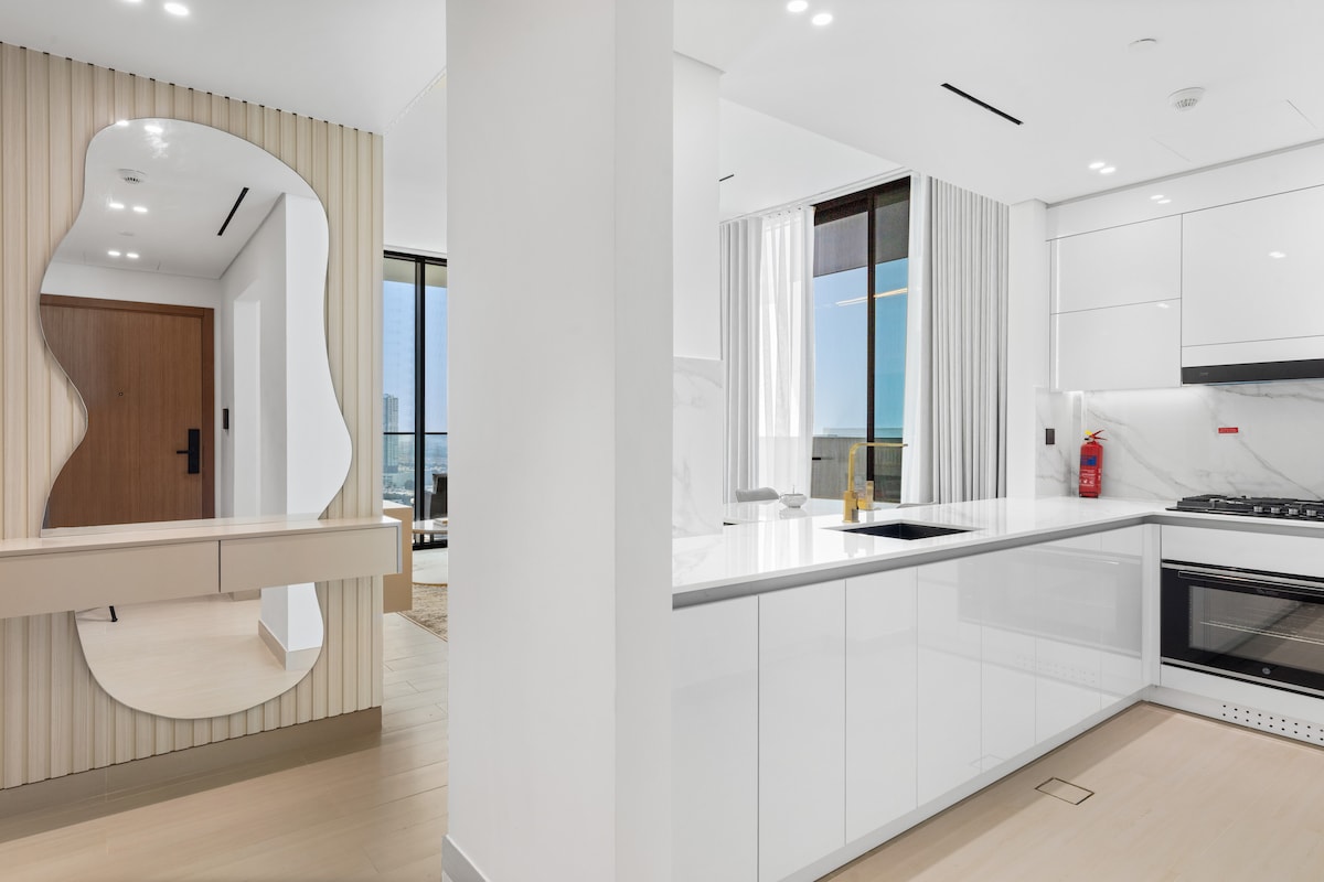 A modern kitchen area showcases sleek white cabinetry and a marble countertop. A dining space is visible, featuring a distinctive mirror reflecting the room. Natural light floods through large windows, highlighting the contemporary design and clean lines of the apartment.