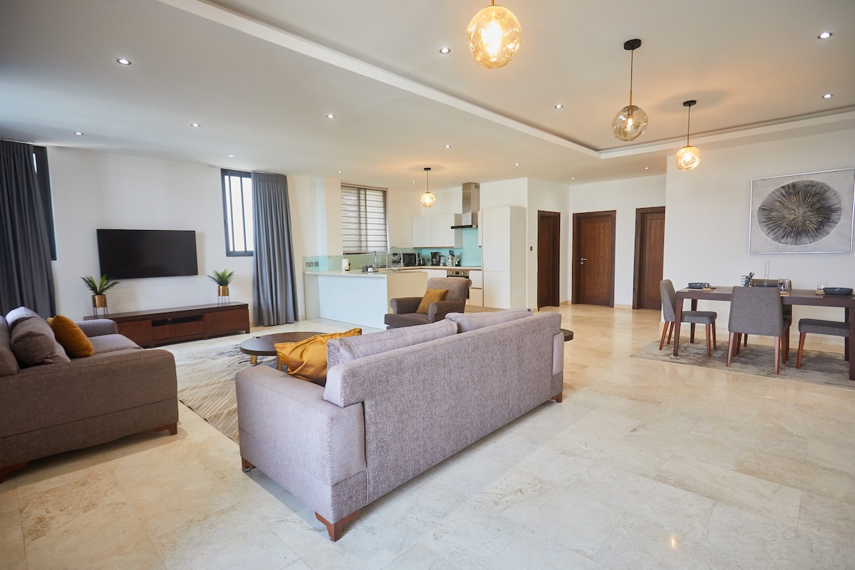 A spacious living area showcases two comfortable sofas in neutral tones, paired with a stylish coffee table. The open layout connects to a well-equipped kitchen and dining area, which is illuminated by modern pendant lights. Large windows allow natural light while maintaining a serene ambiance.