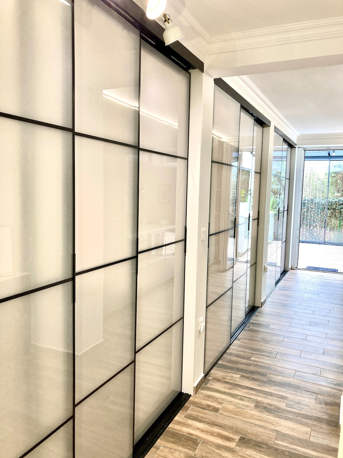 A corridor features floor-to-ceiling sliding doors framed in black, allowing natural light to filter through. Light wooden flooring runs the length of the space, which is complemented by a bright entryway leading to the outside. The walls are adorned with a soft neutral color.