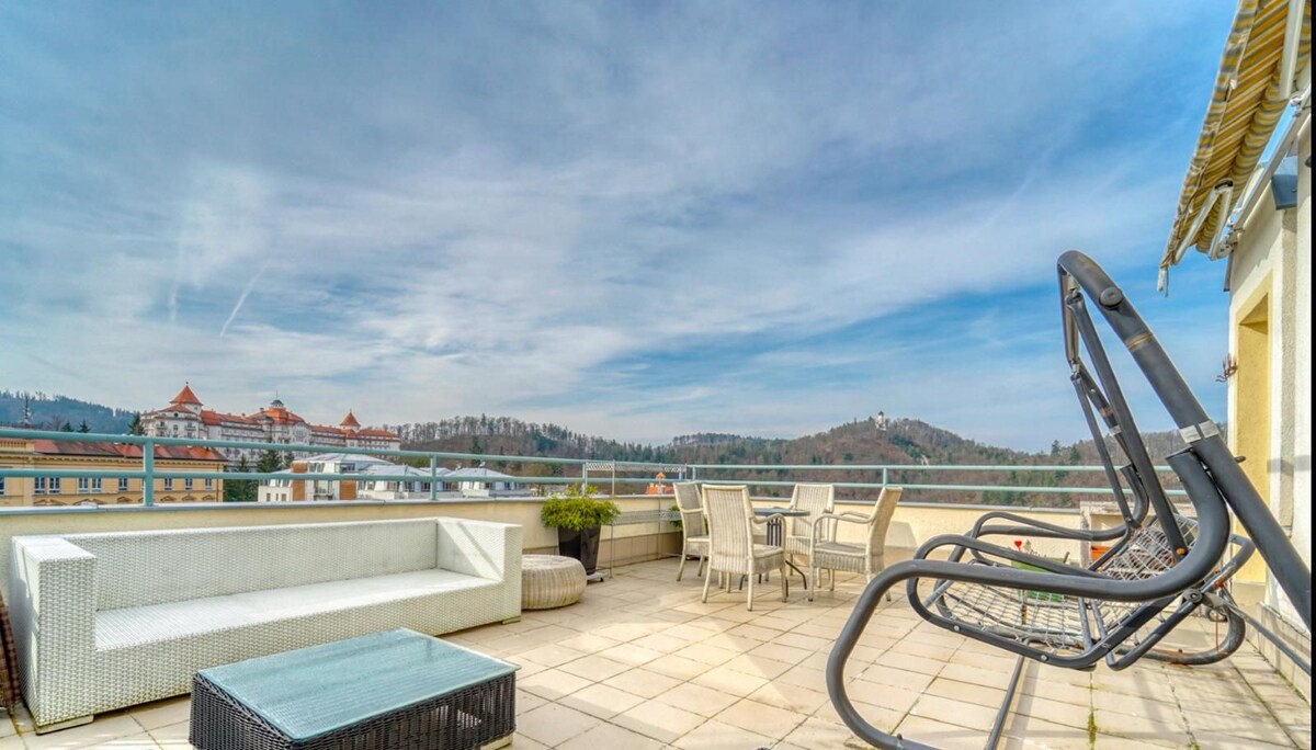 A spacious terrace features comfortable outdoor seating, including a lounge set and a dining table. The surrounding view showcases distant hills and architecture, creating an inviting space for relaxation.