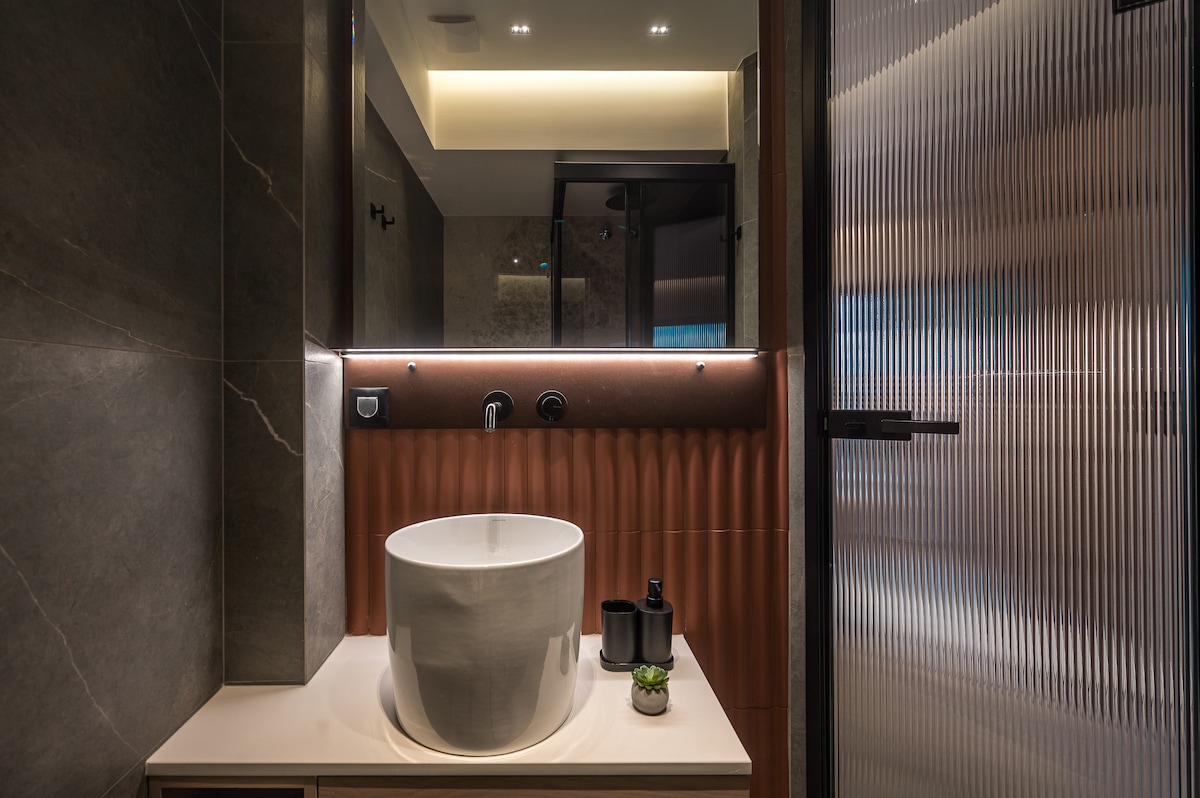 A modern bathroom is highlighted by a circular sink atop a sleek countertop. A glass shower enclosure features textured panels, while ambient lighting accents the space. Dark tiles create a sophisticated backdrop, complemented by minimalist decor, including a small plant and neatly arranged toiletries.