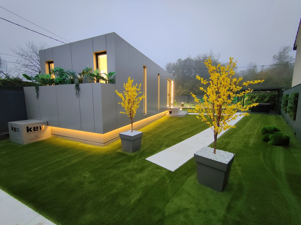 A modern residence is surrounded by well-maintained greenery. Two potted yellow trees add a pop of color, contrasting with the sleek exterior. Pathways are lined with a light surface leading to the structure, which is softly illuminated, enhancing the inviting outdoor ambiance.
