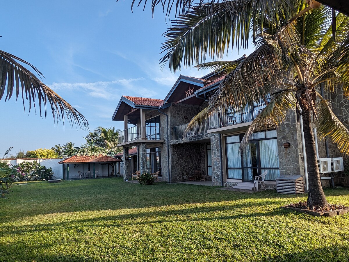 A spacious hotel exterior showcases a two-story stone structure flanked by palm trees and lush green grass. Balconies are visible on the upper level, and a red-roofed gazebo is nestled in the background, creating a serene coastal ambiance.
