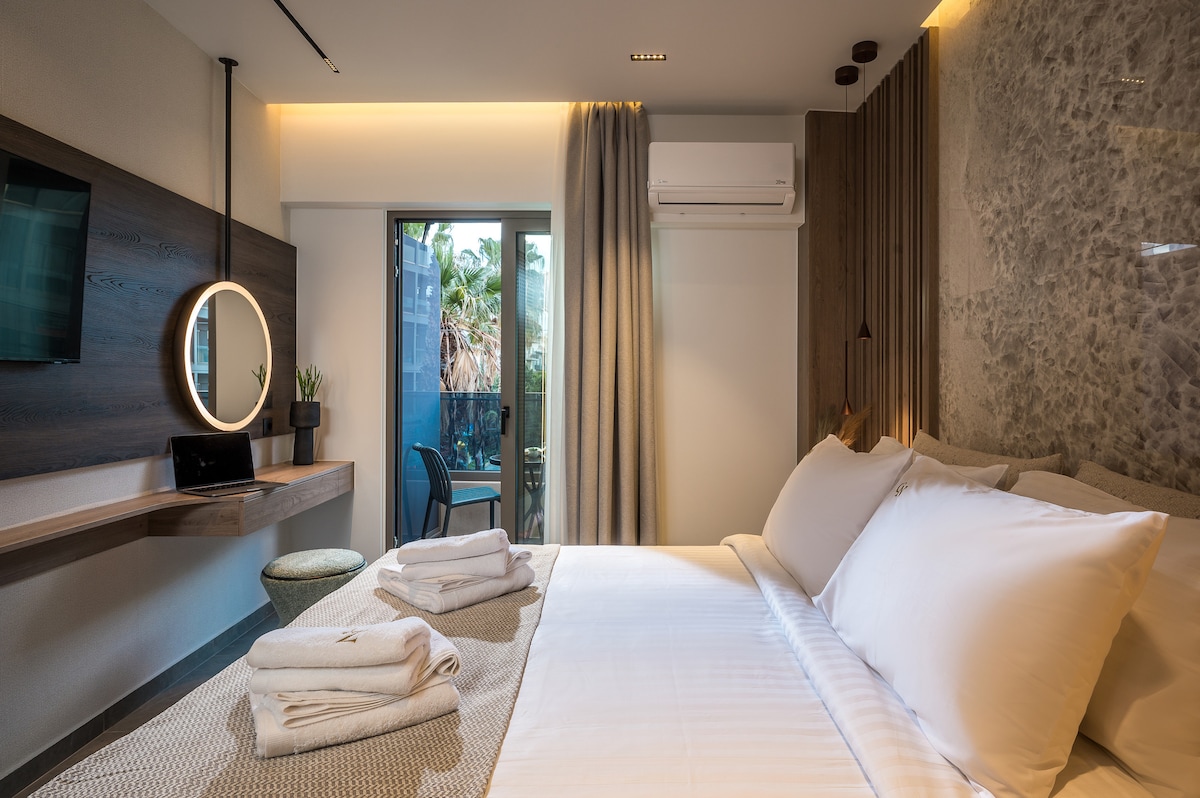 A modern sleeping area features a spacious bed with fresh linens, accompanied by neatly folded towels. A round mirror and a desk are positioned against the wall, while a window allows natural light, offering a view of greenery. Air conditioning is also visible.