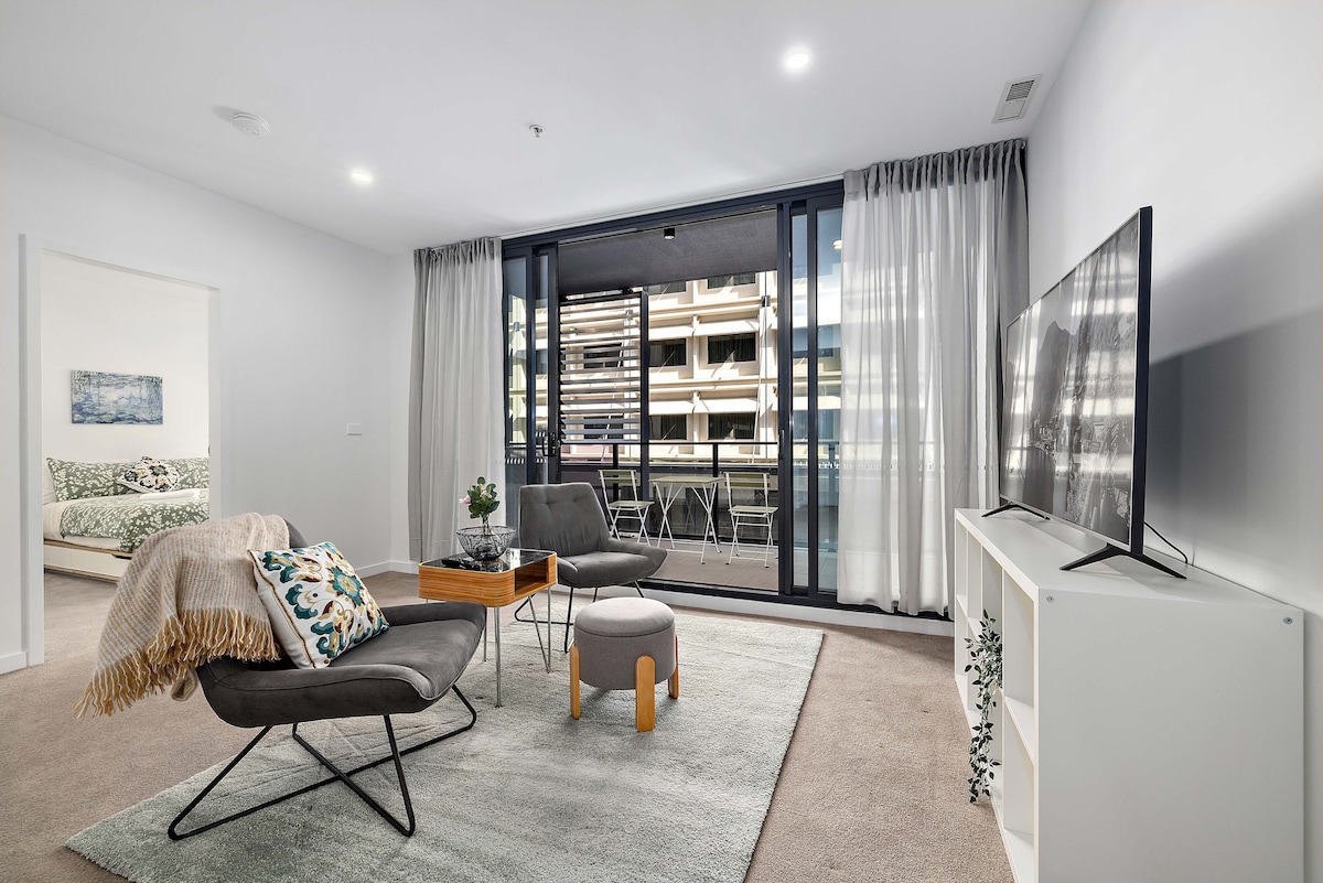 A stylish living area is highlighted by modern furnishings, including a comfortable armchair and a cozy rug. Large floor-to-ceiling windows are adorned with sheer curtains, allowing natural light to fill the space while providing views of an outdoor balcony with seating.