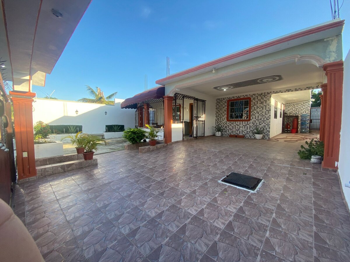 A well-lit outdoor area features a tiled surface and is shaded by an awning. Potted plants accentuate the space, while a pathway leads to a garden area. Two entrance points can be seen, alongside a decorative wall with patterned tiles.