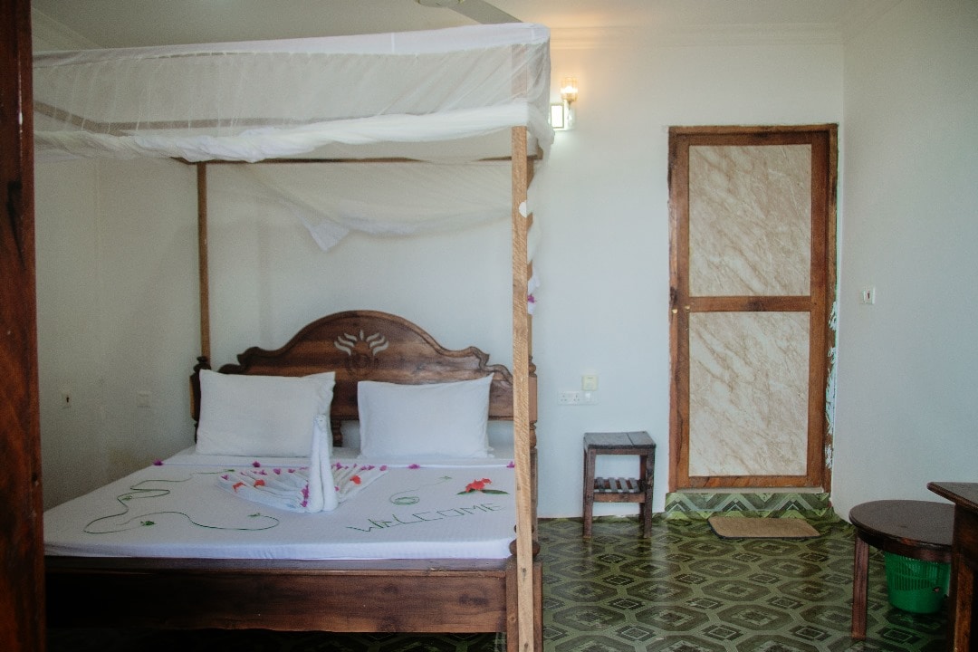 A serene bedroom is highlighted by a wooden canopy bed with white bedding. Decorative petals are arranged on the bed. A small table and a chair are positioned nearby, while a door with natural wooden accents adds a welcoming touch to the space.