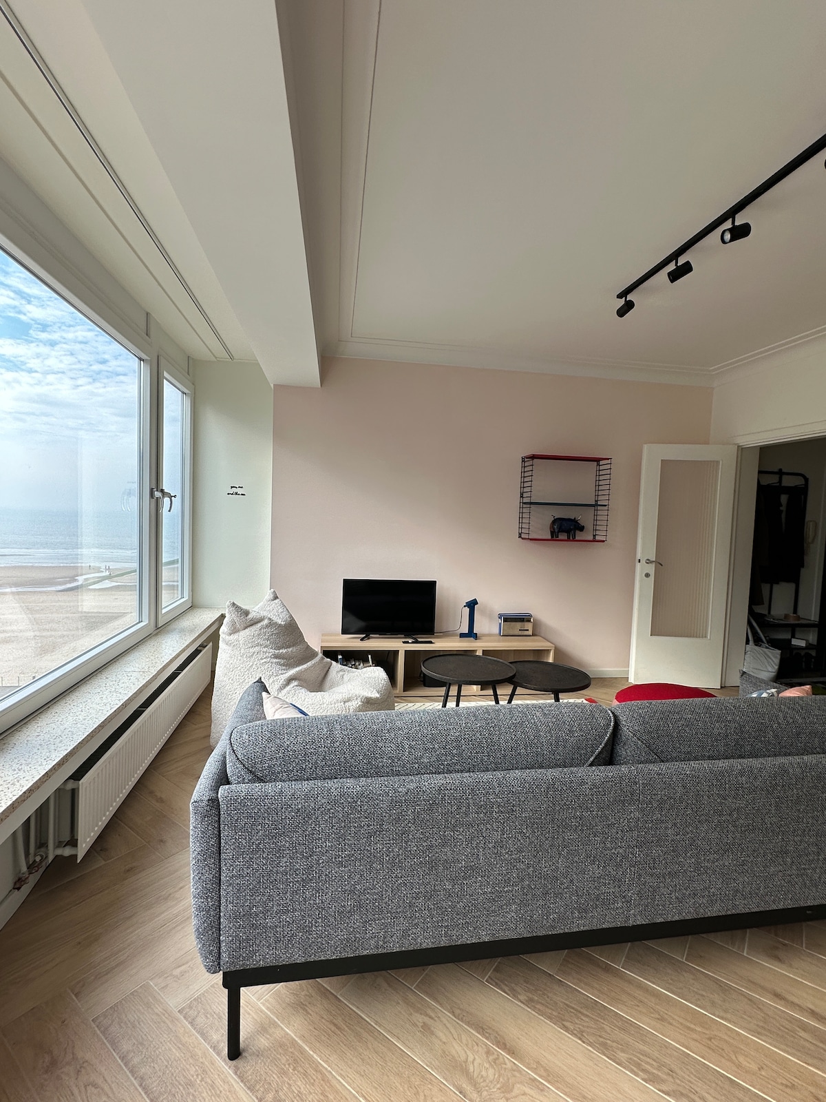 A modern living area features a gray sofa facing a coffee table and a television. Large windows provide an unobstructed view of the sea, allowing natural light to fill the space. Soft textures in the decor create a comfortable ambiance.